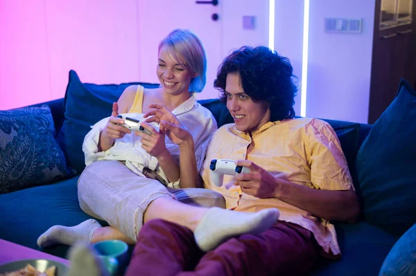 Happy young people playing video games on console while sitting on couch in front of tv. Millennial couple spending fun time together at home. Room with neon lights. — Stock Photo, Image