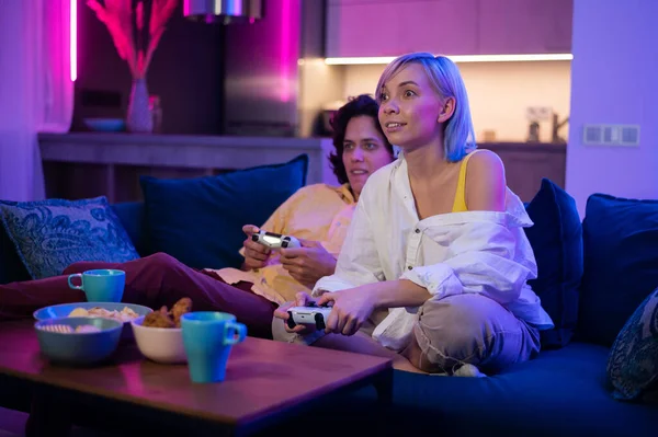 Glückliche junge Leute, die Videospiele auf der Konsole spielen, während sie auf der Couch vor dem Fernseher sitzen. Millennial-Paar verbringt lustige Zeit zusammen zu Hause. Zimmer mit Neonlicht. — Stockfoto