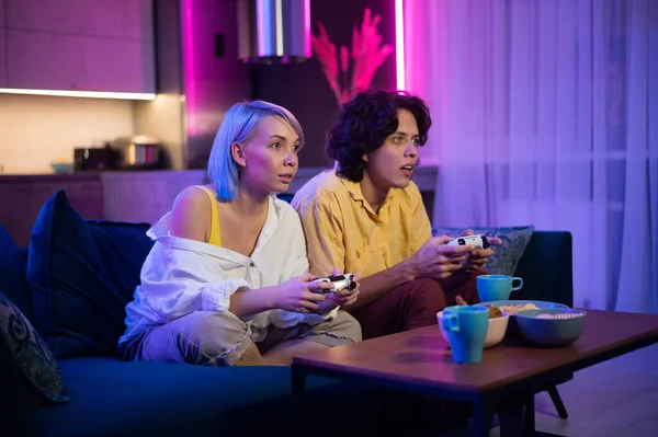 Pareja joven jugando videojuegos mientras está sentada en el piso de la sala de estar iluminada con color neón. — Foto de Stock