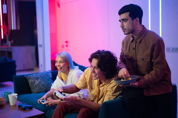 Dos amigos masculinos emocionados por jugar videojuegos en casa mientras una chica se sienta con ellos en un sofá y usa un teléfono inteligente móvil. — Foto de Stock