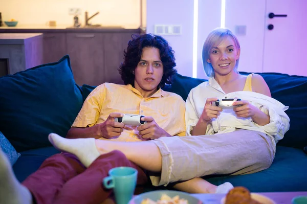 Glücklicher Freund und Freundin, die Videospiele spielen und Steuerknüppel zu Hause halten. Millennial-Paar genießt Spiel, während es auf dem Sofa sitzt und seine Freizeit verbringt. Beziehungskonzept — Stockfoto