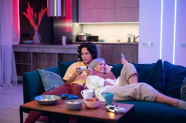 Happy boyfriend and girlfriend laughing after playing video games at home, joysticks and snacks on a table. Millennial couple enjoying spending time together. Concept of relationship. — Stock Photo, Image