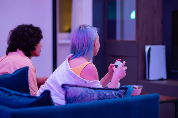 Jóvenes felices jugando videojuegos en la consola mientras están sentados en el sofá frente a la televisión. Pareja milenaria pasando tiempo divertido juntos en casa. Habitación con luces de neón. — Foto de Stock