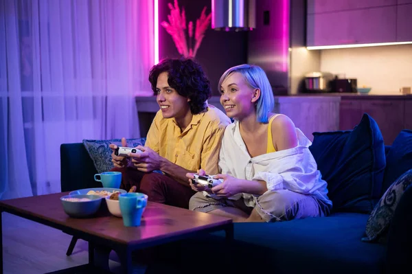 Jóvenes felices jugando videojuegos en la consola mientras están sentados en el sofá frente a la televisión. Pareja milenaria pasando tiempo divertido juntos en casa. Habitación con luces de neón. — Foto de Stock