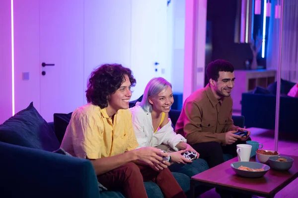 De jeunes amis jouent à des jeux vidéo assis sur un canapé dans un salon. — Photo