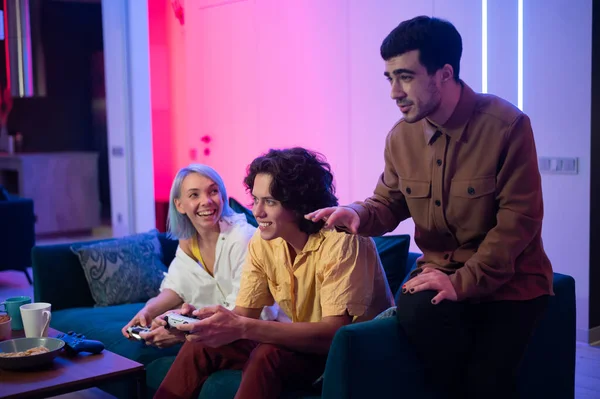 Jeunes gens heureux jouant à des jeux vidéo sur la console tout en étant assis sur le canapé devant la télévision. L'homme conseille son ami dans un jeu. Chambre avec lumières chaudes et néons. — Photo