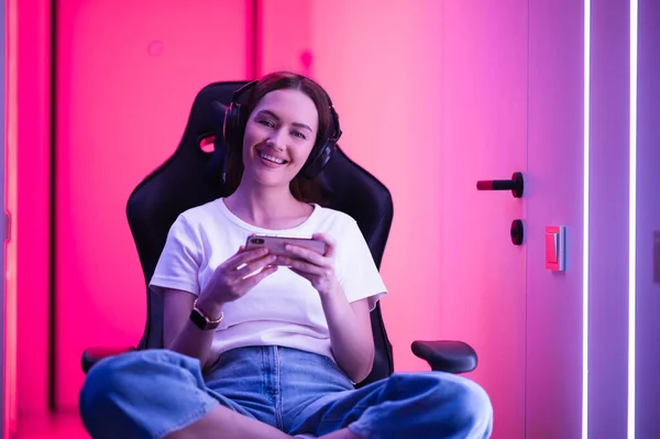 Gamer Girl in einem großen professionellen Musik-Kopfhörer in weißem T-Shirt und blauer Jeans hält Smartphone in der Hand und spielt Spiele-App auf Handy, lächelt und schaut in Kamera drinnen. — Stockfoto