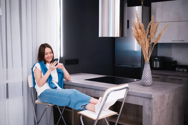 Compras o pedidos de entrega en línea con teléfono inteligente móvil. Mujer joven feliz usando el teléfono celular en casa mientras está sentado en una cocina. Imagen de archivo