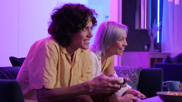 Ung vuxen bror och syster spelar tv-spel sitter i ett rum upplyst med neon färg hemma. — Stockvideo