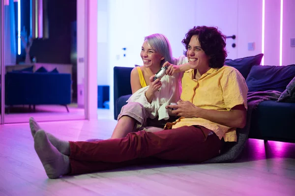 Jeunes gens heureux jouant à des jeux vidéo sur la console tout en étant assis sur le canapé devant la télévision. Couple millénaire passer du temps amusant ensemble à la maison. Chambre avec néons. — Photo