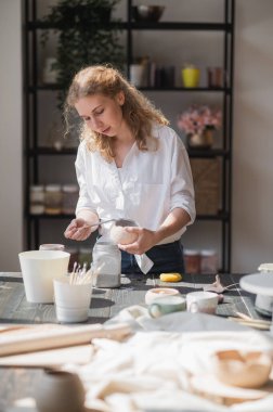 Genç Avrupalı güzel bir çömlekçi atölyedeki masada seramik yapma sürecinde. Güneşli bir günde çömlek atölyesinde saçlarım rüzgarda uçuşuyor..
