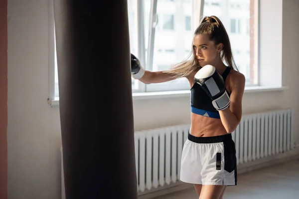 Una chica en ropa deportiva se dedica al boxeo y resuelve un golpe con la mano en un saco de boxeo — Foto de Stock