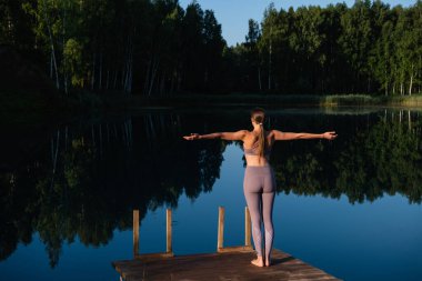 Genç bir kadın orman göl manzarasının üzerinde tahta iskelede yürüyor, doğada sabah yoga çalışmasına hazırlanıyor..