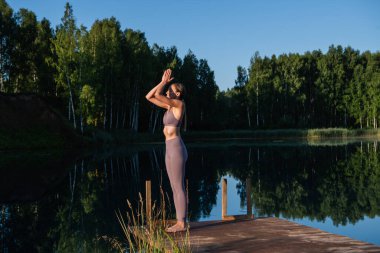 Gölün kenarında yoga yapan genç bir kadın ahşap platformda hareket ediyor. Doğada egzersiz yapan güzel bir genç kadın, sağlıklı bir yaşam tarzı genç insanlar pozitif titreşimler alıyor..