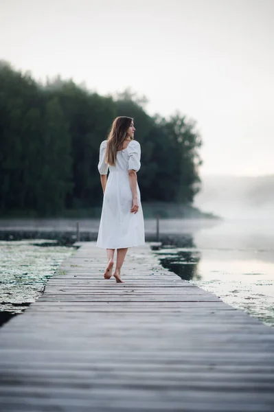 Вид сзади на молодую женщину в белом платье, стоящую в одиночестве на пешеходном мосту и смотрящую на озеро. Туманное холодное утро с туманом над водой. — стоковое фото