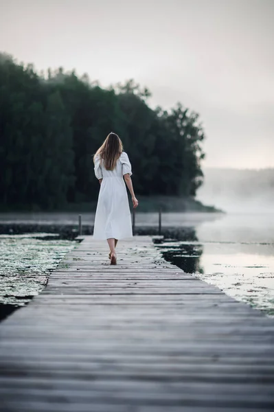 Женщина в белом светлом платье бежит по пирсу на туманном утреннем озере. — стоковое фото