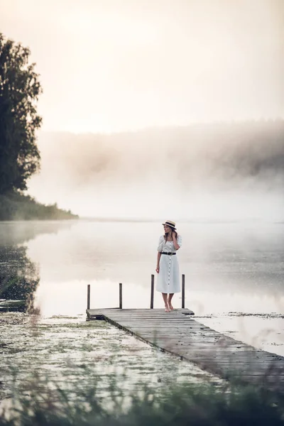 Frau in weißem Kleid und Hut steht auf einem hölzernen Steg mit nebligem See bei Sonnenaufgang. — Stockfoto