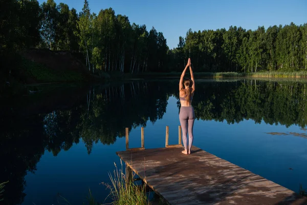 Молода жінка практикує йогу на дерев'яному причалі біля озера. Єдиний вид спорту здорове тренування на природі в сонячну погоду — стокове фото