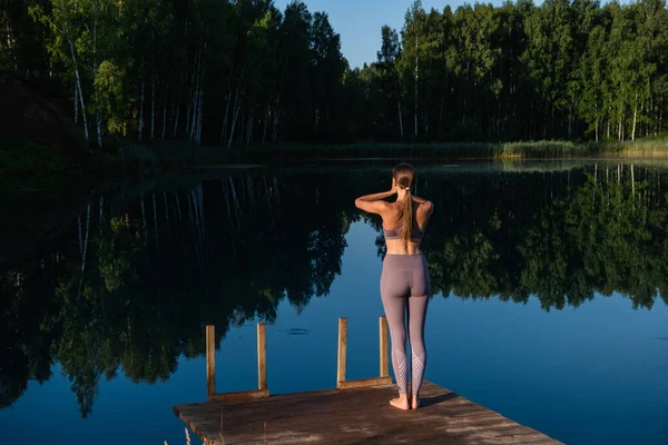 Giovane donna cammina su un molo di legno sopra paesaggio lago foresta, incrocia le braccia in un gesto namaste. Donna braccia tese in natura. — Foto Stock