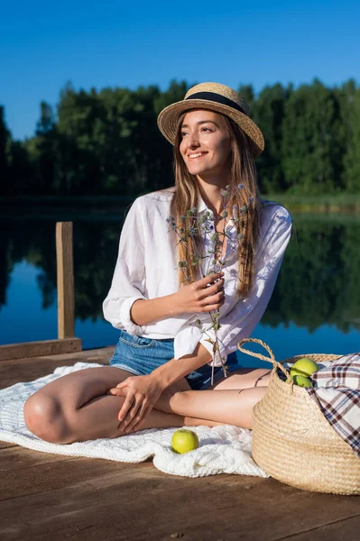 Beautiful young woman wearing blue shorts, shirt and hat enjoys her morning picnic on a wooden pier near the lake. — Φωτογραφία Αρχείου