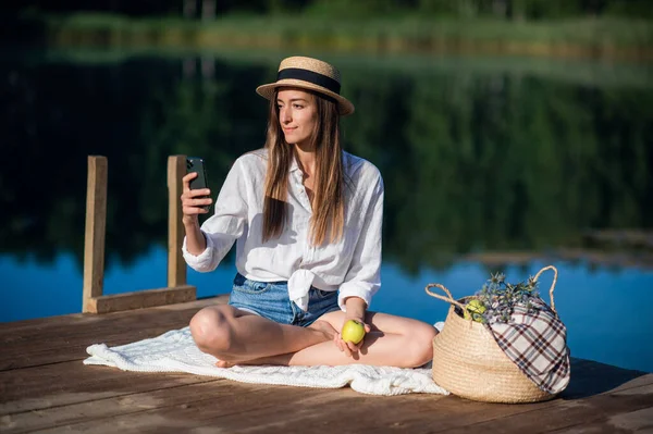 모자를 쓴 아름다운 젊은 여자가 여름 숲 근처 호수 근처에서 피크닉을 한다. 사과를 먹으면서 스마트폰을 보고 있어요. — 스톡 사진