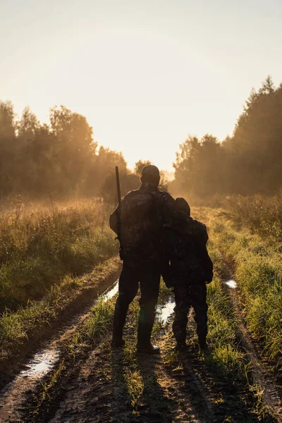 Caçadores com equipamentos de caça que vão embora através do campo rural em direção à floresta ao pôr do sol durante a temporada de caça no campo. — Fotografia de Stock