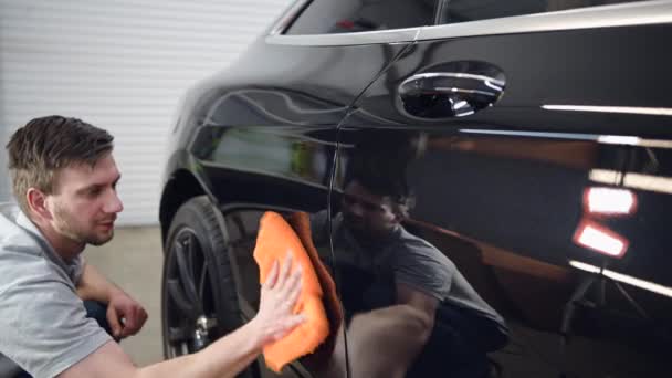 Secagem de carro. trabalhador masculino com pano de laranja limpa gotas de água da porta do carro após a lavagem do carro. — Vídeo de Stock