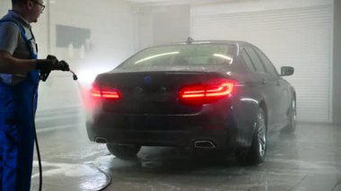 Adam araba tamirhanesinde araba yıkamak için yüksek basınçlı çamaşır makinesinden su püskürtüyor..