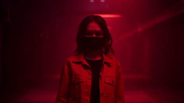 Γυναίκα στέκεται φορώντας προστατευτική μάσκα προσώπου σε ένα κόκκινο φως της σκοτεινής υπόγειας σήραγγας και κοιτάζοντας κάμερα. Covid-19 πανδημία - κατάθλιψη και θλίψη. — Αρχείο Βίντεο