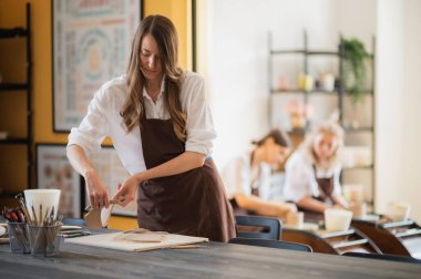Önlüklü üç kadın çömlekçilikte çalışıyor, kil yoğuruyor ve atölyede toprak eşyalar yapıyor.