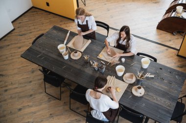 Üç önlüklü kadın masada oturuyor, killeri yoğuruyor ve derste toprak eşyalar yapıyor.
