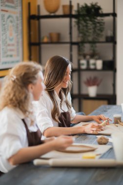 Önlüklü iki kadın masada oturuyor, killeri yoğuruyor ve derste toprak eşyalar yapıyor.