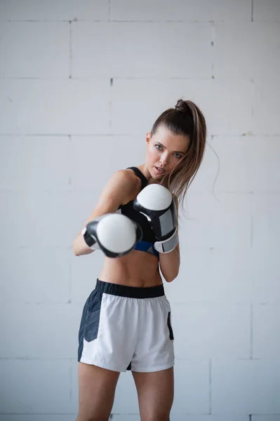 Una bella atleta donna in guanti da boxe e uniforme sportiva si erge contro un muro di mattoni bianchi — Foto Stock