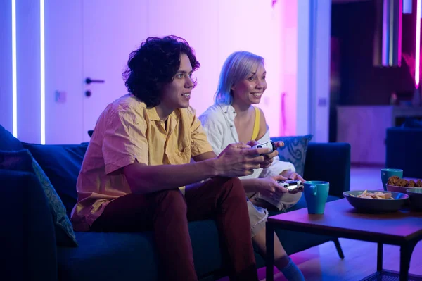 Pareja sonriente sosteniendo mandos jugando videojuegos en casa. Inyección de bajo ángulo de los jóvenes que pasan tiempo juntos durante el aislamiento de sí mismos en pandemia. Imagen de archivo