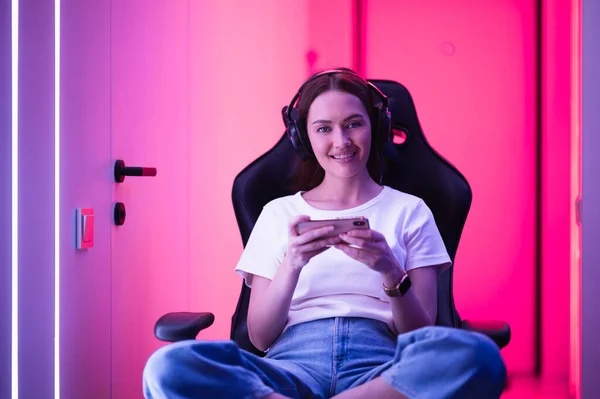 Junges Mädchen spielt mobiles Online-Spiel auf einem Smartphone, während es auf einem Spielstuhl in einem bunten Neonlicht-Raum sitzt. — Stockfoto