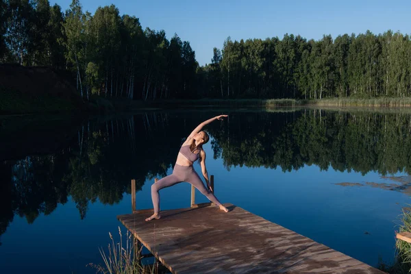 Egy nő jógázik egy erdei tavon. Gyönyörű lány csinál gyakorlatokat nyújtás és rugalmasság szabadban — Stock Fotó