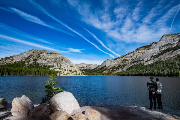 Pareja joven disfruta de vistas al lago Tenaya en Yosemite, EE.UU.: Paisaje de montaña con lago espejo rodeado de picos altos — Foto de Stock