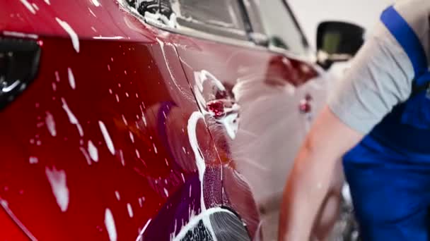 Hombre trabajador lavando coche rojo en un lavado de coche con guante negro. — Vídeos de Stock