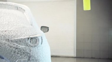 İşçi garajda yıkanmak için sabun köpüğüyle araba gövdesini kaplıyor, hortumla spreyliyor, araba yıkıyor..