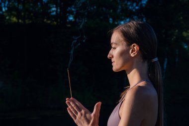 Yoga yapan ve güneşin doğuşunda meditasyon yapan bir kadın ellerinde aroma çubuğu, ellerinde namaste jesti olan bir kadın..