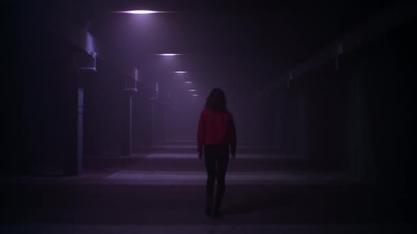 Camera follows young brunette woman in red jacket walks quickly along long corridor with flashes in the dark. — Stock Video