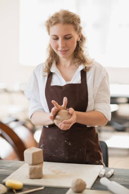 Kadın usta çömlek atölyesindeki ahşap masanın yanında kil yoğuruyor.