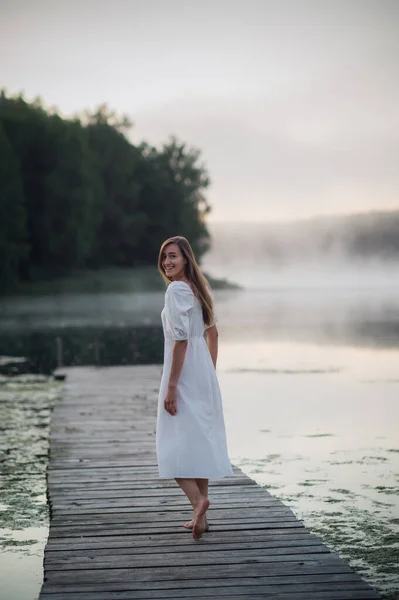 Vista posterior de la mujer joven en vestido blanco de pie solo en la pasarela y mirando al lago. Niebla mañana fría con una niebla sobre el agua. Fotos de stock libres de derechos