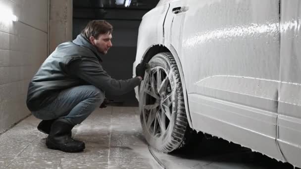 Espuma gotas de un SUV moderno durante el lavado de coches. Hombre usando cepillo para limpiar la superficie de la llanta de la rueda de un coche en el lavado automático — Vídeos de Stock