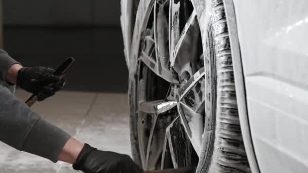 Manos de hombre joven sosteniendo cepillo de limpieza especial, lavando la rueda del coche con espuma. Limpieza de llantas modernas de coches de lujo en el servicio de lavado de automóviles. — Vídeos de Stock