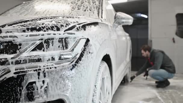 Espuma gotas de un SUV moderno durante el lavado de coches. Hombre usando cepillo para limpiar la superficie de la llanta de la rueda de un coche en el lavado automático — Vídeos de Stock