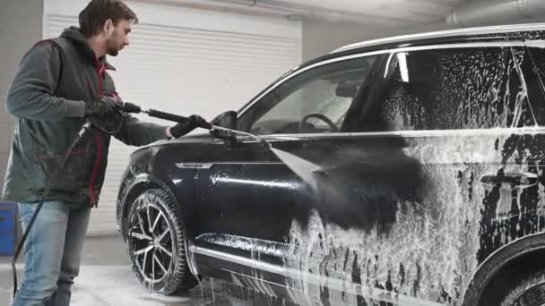 Biały mężczyzna pracuje w myjni samochodowej, a pracownik myjni myje samochód. Pracownik serwisu myje pianę z maszyny silnym ciśnieniem wody. — Wideo stockowe