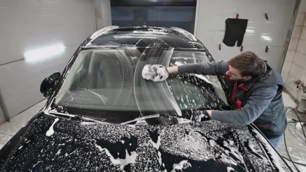 Hombre trabajador de limpieza de la ventana delantera de un coche cubierto de espuma. — Vídeos de Stock