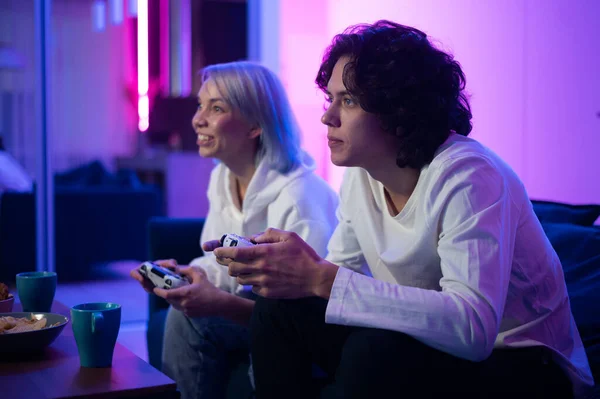 Двое молодых людей играют в видеоигры дома. Эмоциональные разноплановые геймеры, держащие джойстики и конкурирующие в интенсивной видеоигре на игровой консоли. — стоковое фото
