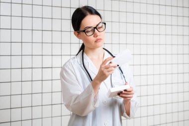 Genç, zayıf, koyu renk saçlı, gözlüklü bir kız, tıbbi bir kıyafet giymiş, eczanenin arka planındaki ilaç paketinin yanında bir şeyler okuyor..